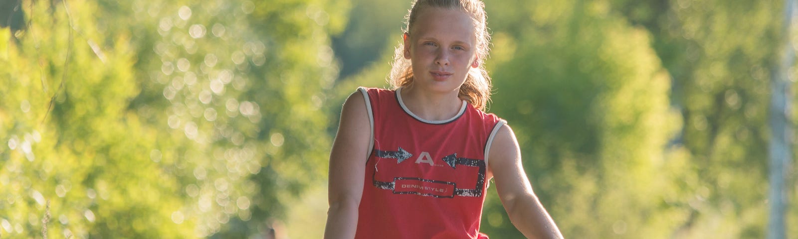 Young girl with a ponytail in a red tank top, sitting on a bicycle on a country path.