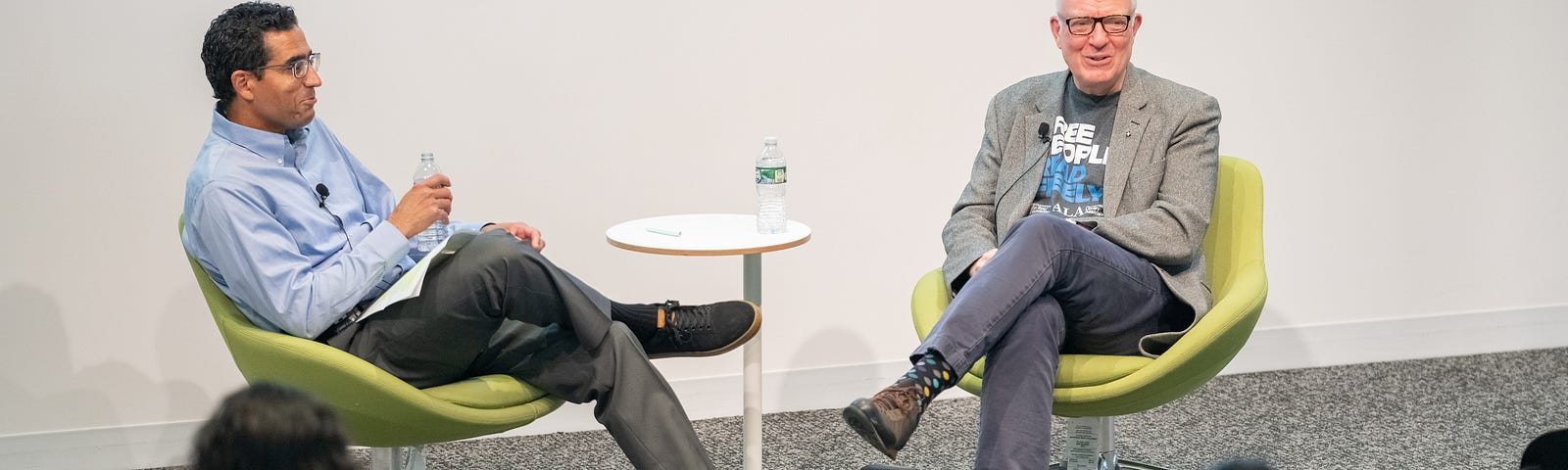 Photo of two men in business casual attire sitting in two chairs in front of a crowd.