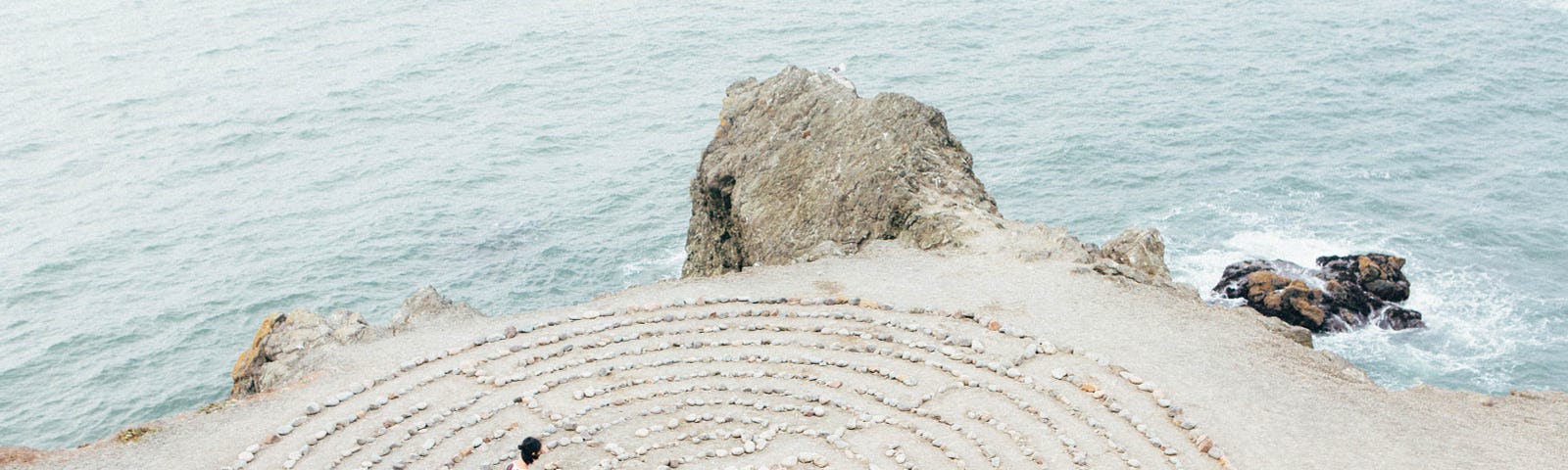 A person walks by the sea, amongst pebbles arranged in a criuclar shape