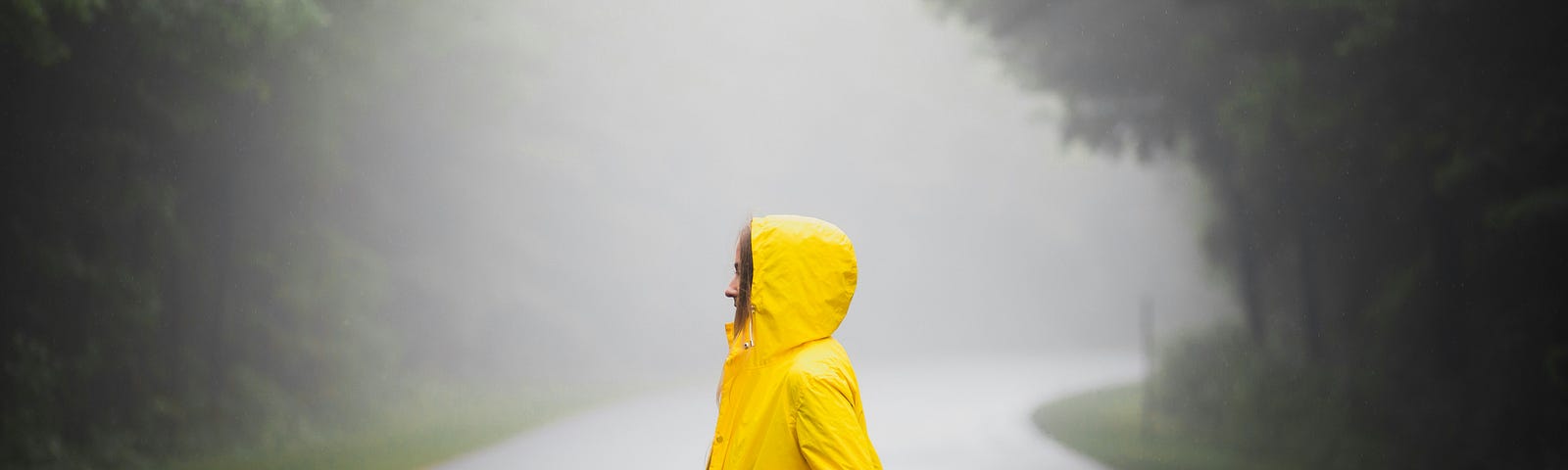 A woman standing in a yellow raincoat on a deserted wooded street.