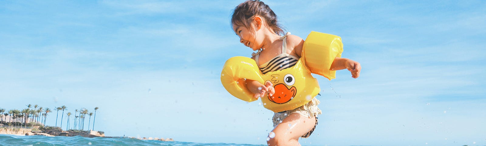 Image shows little girl in striped bathing suit and yellow ducky inflatable life vest/water wings happily playing and splashing on the shallow surf of a beach. Blue sky in the background and palm trees in the distance.