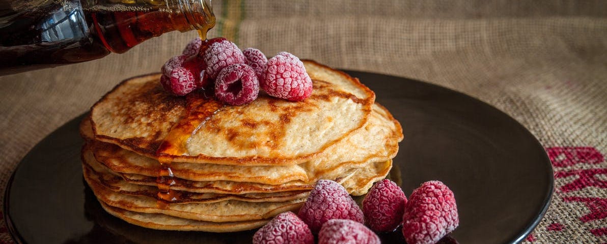 A stack of pancakes and frozen berries on a plate being covered in maple syrup poured from a bottle