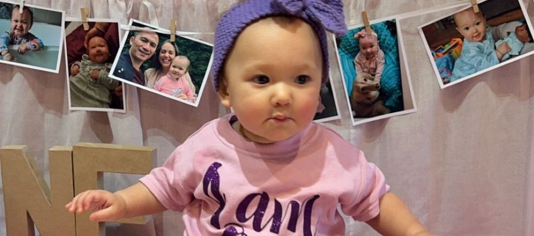 A baby sits on table in front of photos of her and her parents. She’s wearing a top that says “I am one”. A birthday ‘cake’ carved from melons and decorated with strawberries sits on the end of the table.