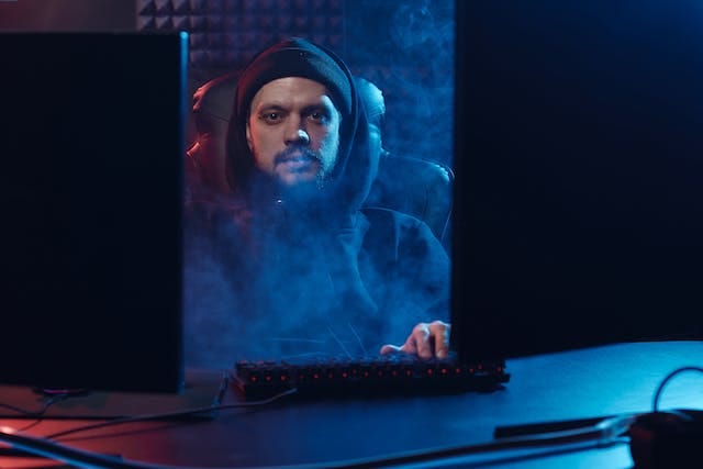 A man in a hoodie sitting between two monitors in a smoky room