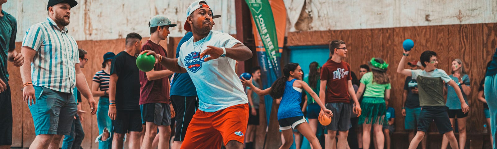 A male adult rears back to throw a dodgeball in a dodgeball game in a gym-like atmosphere.