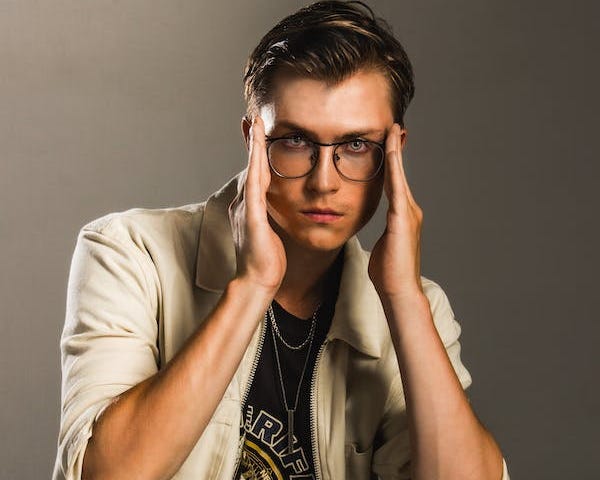 Slender, young man in glasses and trendy clothes looking at the camera in a model pose. He’s neither smiling nor frowning, but instead looks focused. The background a flat grey canvass, like this is in a studio