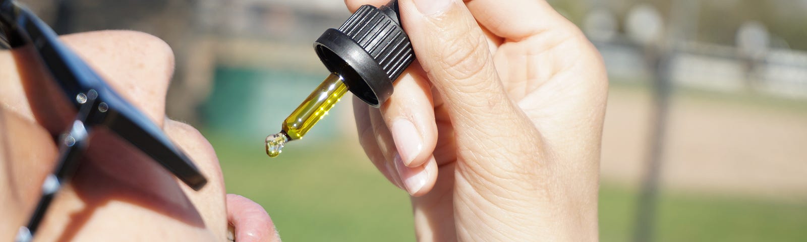 Woman with sunglasses uses an eyedropper to put amber-colored cannabidiol (CBD) into her mouth.