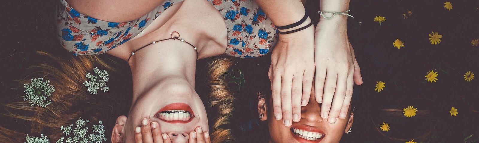 Image shows two women lying on their backs on the grass and holding their hands in front of each other’s eyes. Both are smiling.