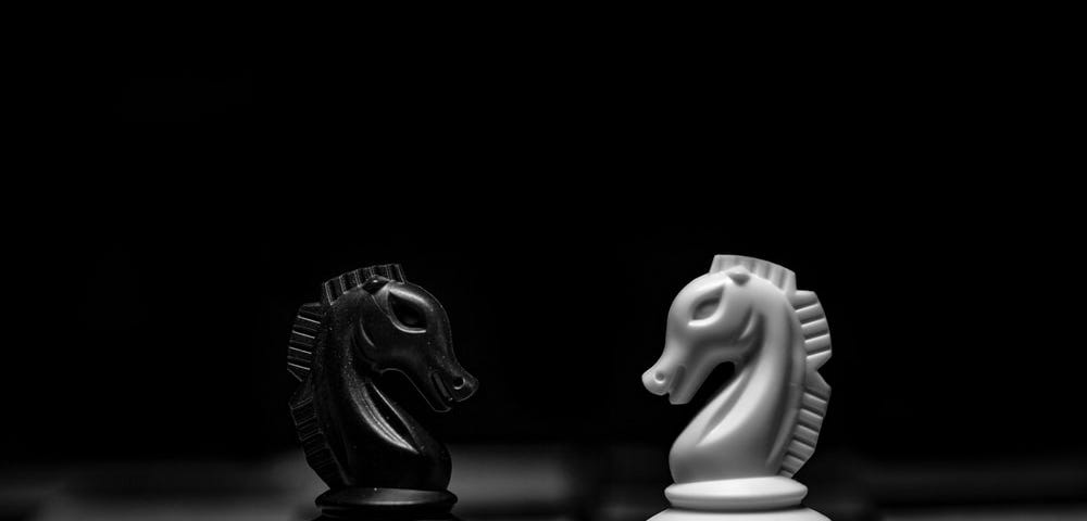 A black and white photo of two chess knights on a chessboard facing each other.