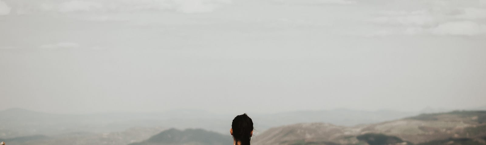 A woman sits on the earth, looking out over a landscape of hills and grey sky.