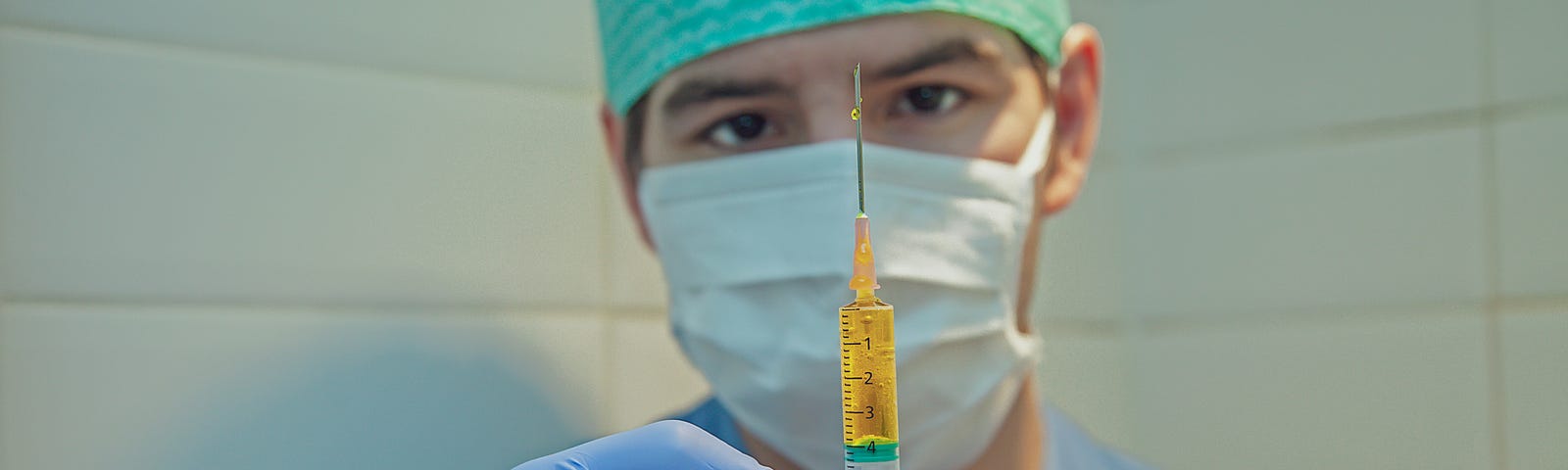 A health care professional, wearing a mask and dressed in scrubs, holds up a syringe, ready to give someone an injection.