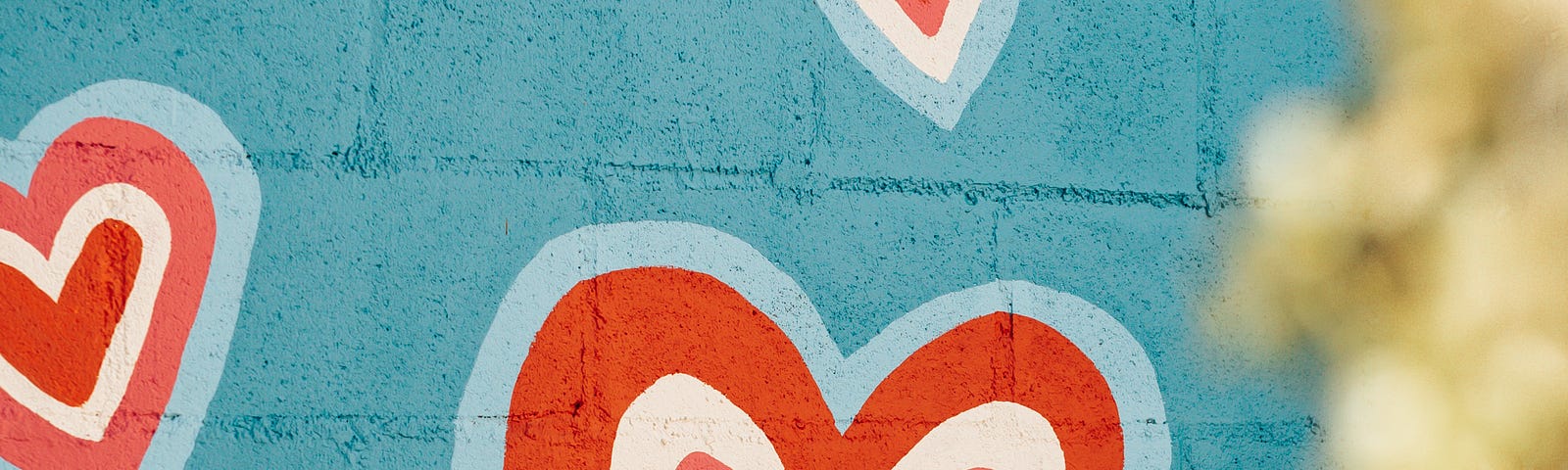 Pink hearts outlined in white, red and light blue painted on a blue cinder block wall.