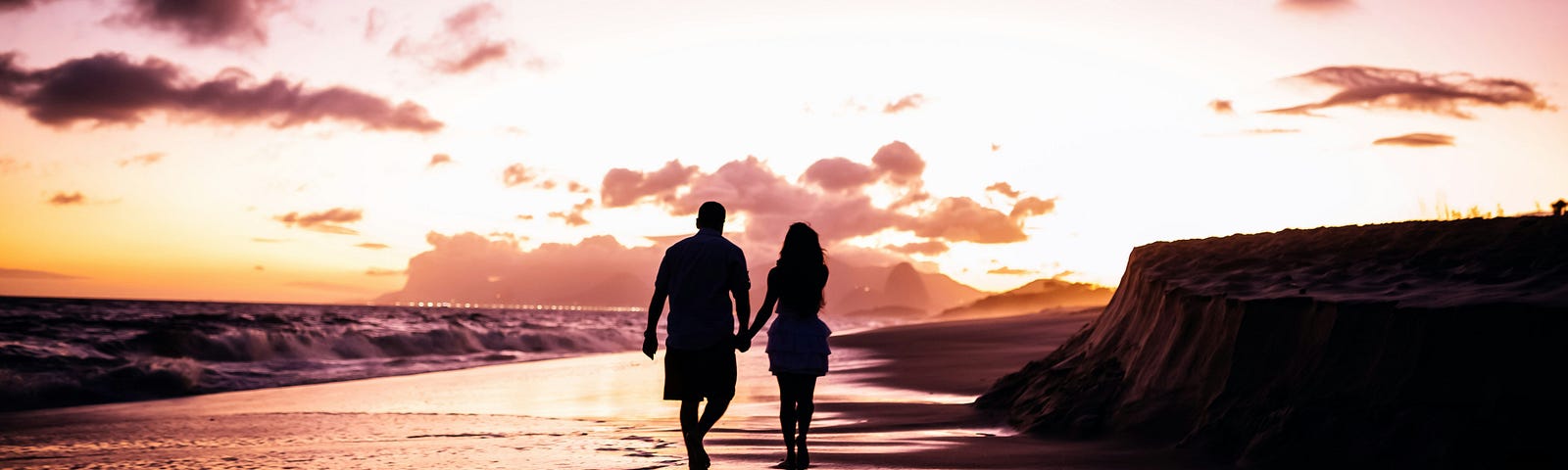 A silhoutted couple walk hand in hand along the waters edge on a sunset beach