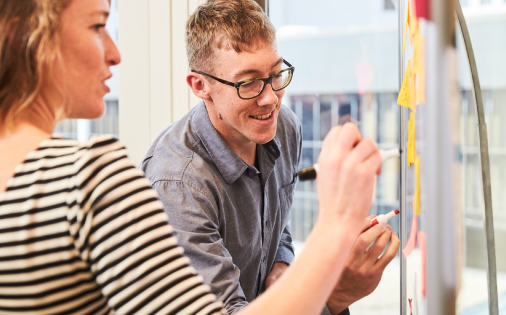 Two colleagues work together at a whiteboard, writing on several posted sticky notes.