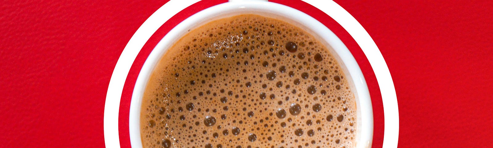 A bird’s eye view of a white coffee cup of frothy coffee on a red background.