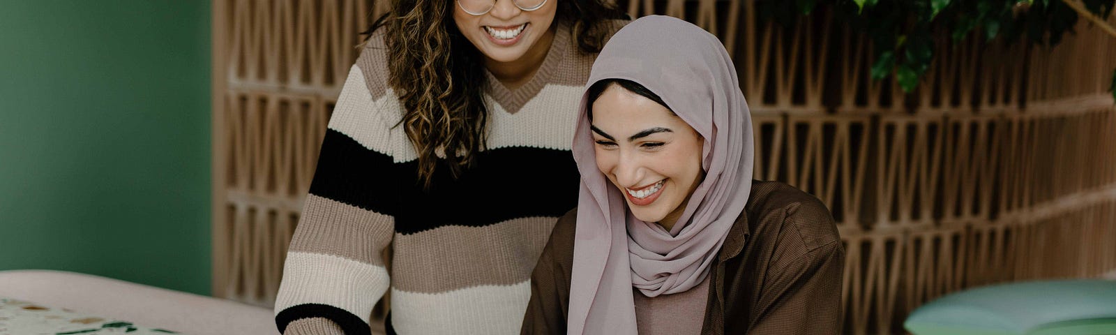 An Asian woman with curly brown hair, left, wears a striped brown, beige, and black sweater. From where she stands, she looks down at a notebook and smiles. A woman in a light purple hijab, right, wears a brown jacket and smiles as she looks at a tablet. The two are at a white table with various color splotches.