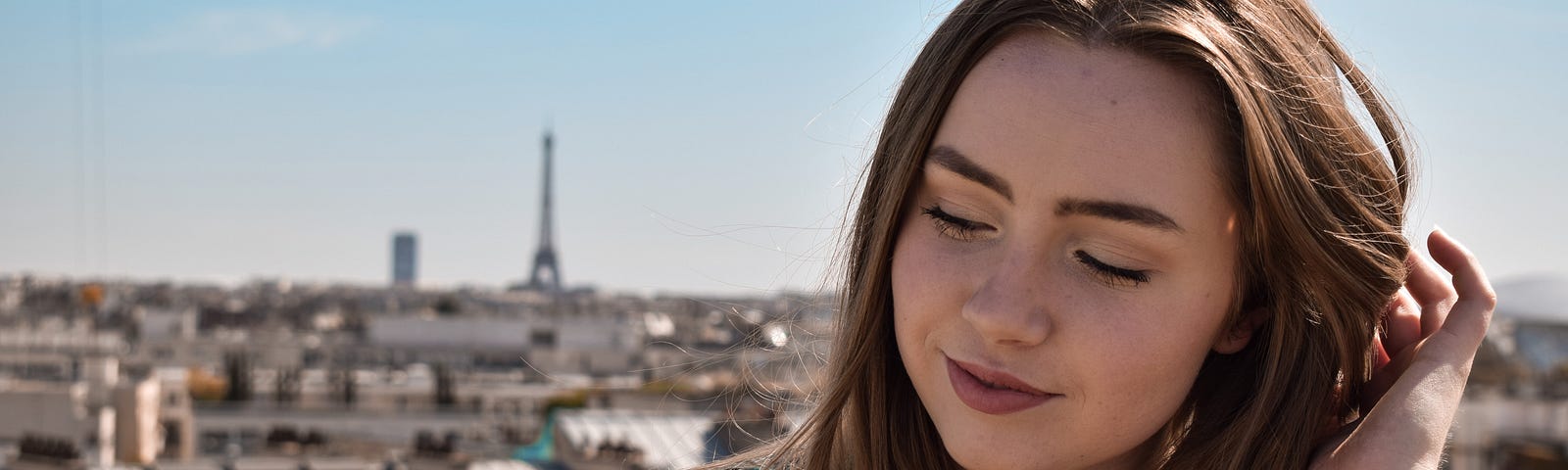 A brown-haired woman scratches her head. The Eiffel Tower can be seen in the distance, against a clear, blue sky.