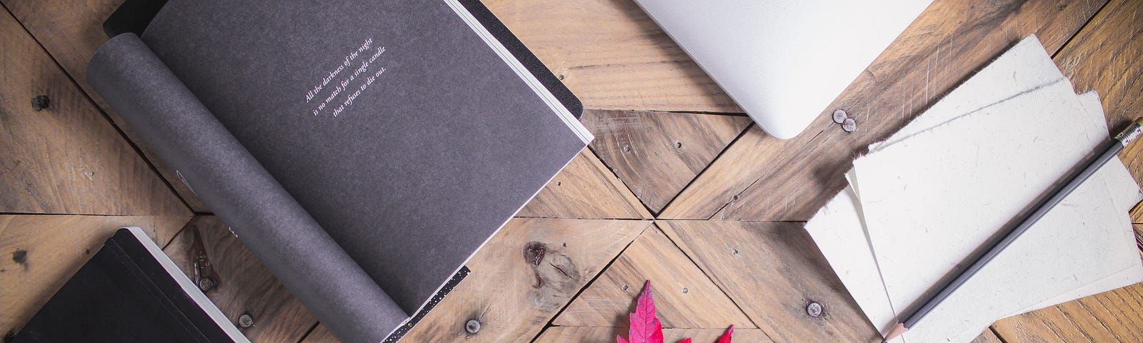 A black journal, a book, a red leaf, paper, a laptop, index cards, and glasses on a light brown wooden table