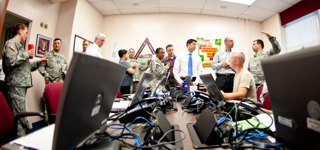 Bilgisayarlar ve kablolarla dolu bir masanın önünde ABD askeri subayları ve sivillerle dolu bir toplantı odası