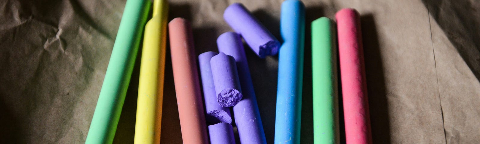 A set of rainbow-coloured chalk displayed on a brown paper. The purple chalk is broken into several pieces.