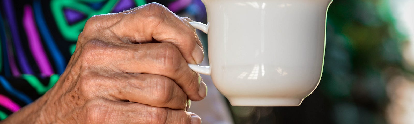 Hands of an elderly lady in a colorful dress and holding a tea cup.