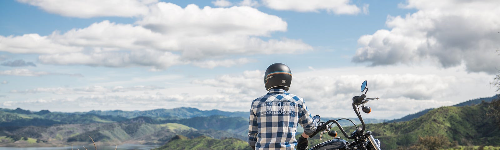 A person wearing a black helmet and a blue and white checked shirt leans against a black motorcycle and looks out at mountains and a river under a blue sky that is dotted with puffy clouds.