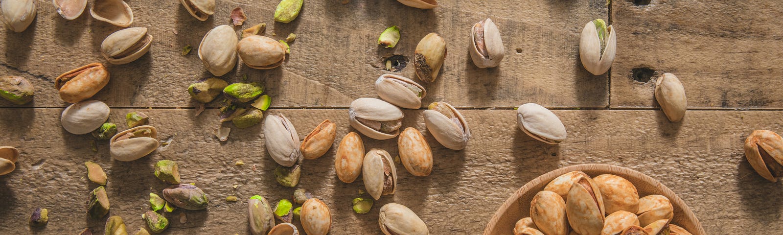 Photo taken from overhead of some pistachios in a little bowl with many others strewn across a wooden table or counter. Photo by Brenan Greene on Unsplash