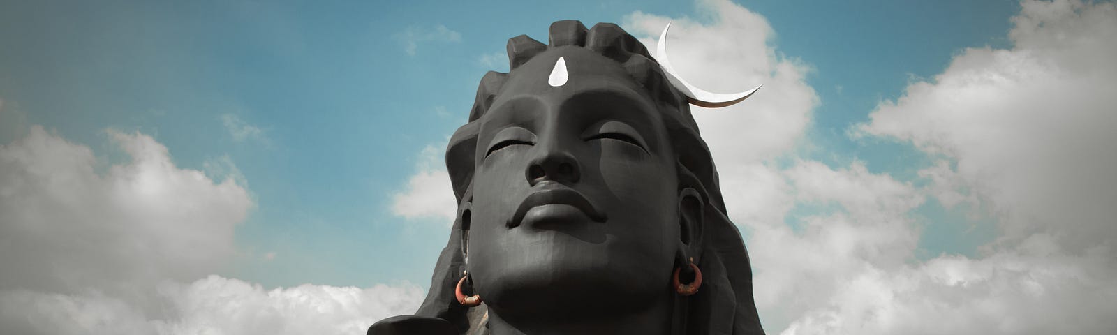 A statue of a long-haired woman in stone from the neck up, eyes closed, against the backdrop of sky and clouds with a sliver of moon touching the right side of her head. The statue is dark gray all over except for her orange-brown hoop earrings and a large white teardrop shape on her forehead.