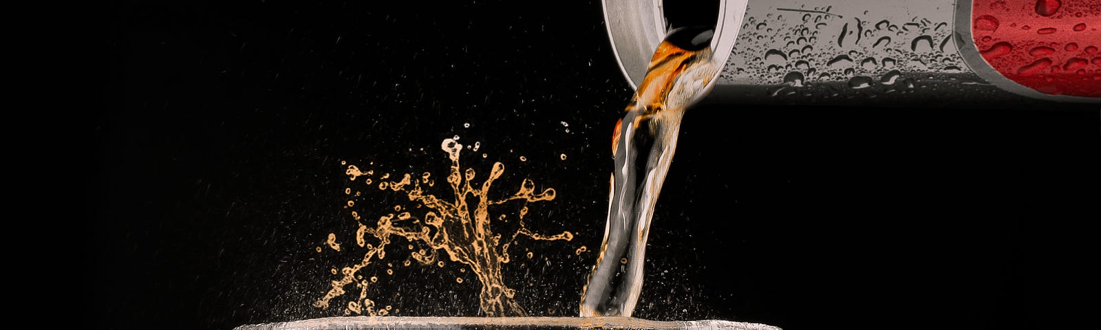 Soda pours from a can of Coke, in the upper right corner of the image. The glass is octagonal in shape, clear, and nearly full of soda. A small amount of the beverage splashes upward out of the glass. Black background.