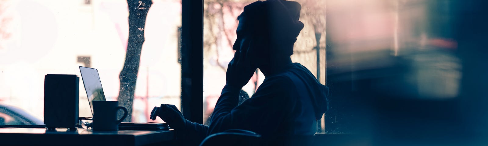 A figure in silhouette sits in a cafe, nursing a mug of coffee or tea. They have a laptop on the table in front of them, and they appear to be thinking through a problem.