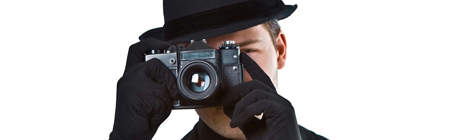 Man in suit, hat and gloves holding a camera in front of his face to take a picture.