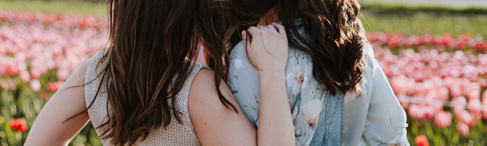 Two women stand with arms around each other facing away from the camera towards a field of flowers.