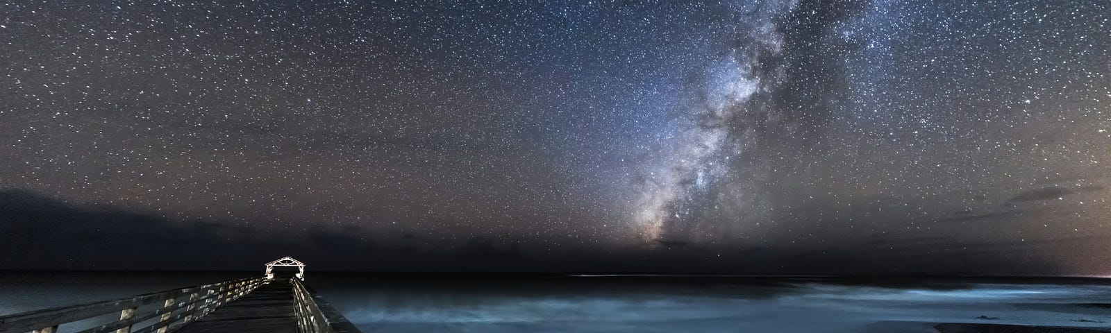 The Milky Way and stars dominate the night sky above a lake, with a pier in the foreground.
