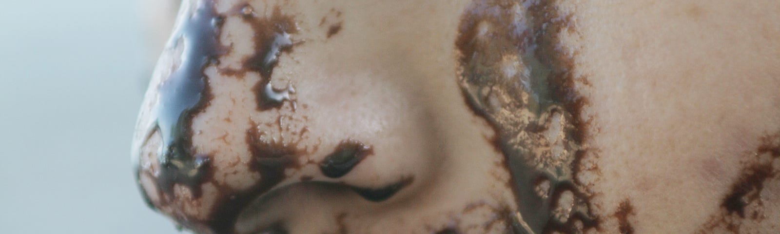 White woman eating chocolate. The dark chocolate is smeared over her lower face. We see her in profile, facing left. Light blue background.