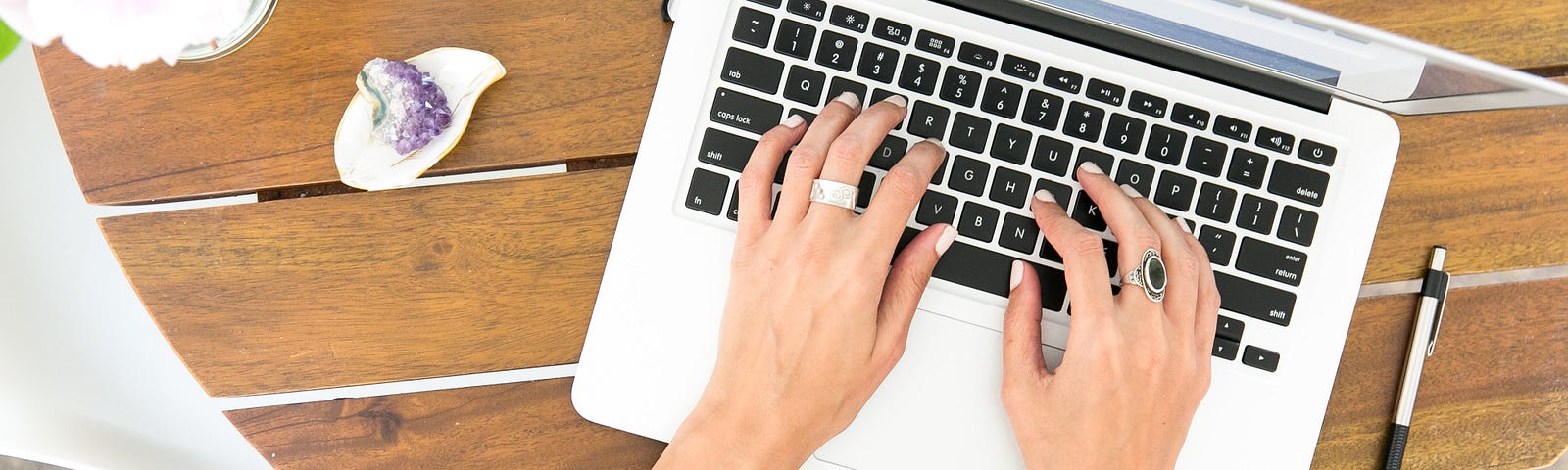 Computer on a planked desk and female hands with rings and bracelets resting on the keys.