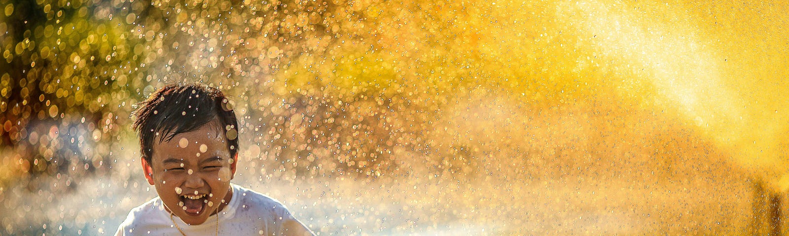 Young boy filled with joy and hope running through a sprinkler of gold lit water. Yellow is the predominant colour.