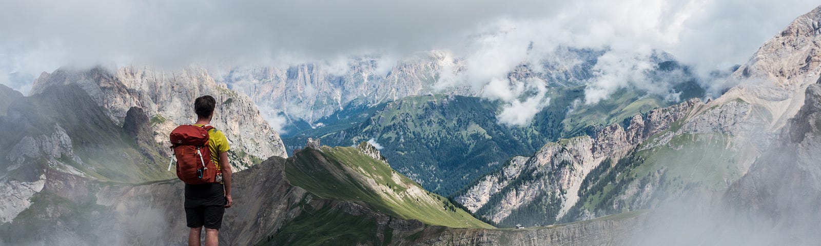 A hiker in a yellow shirt and black short with a red backpack stands on a rocky outcropping looking away from the camera toward a vista of blue, green, and grey mountains.