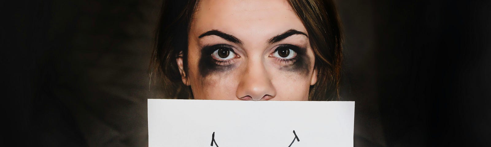 A woman with smeared eye make up, clearly from crying, holds up a piece of paper in front of her mouth with a smile drawn on, trying to hide the truth of how she is feeling.