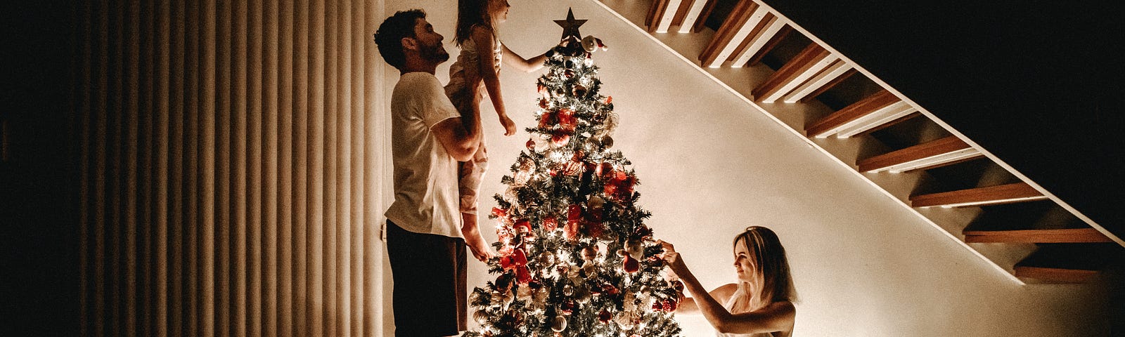 A family decorates a christmas tree. A man holds up a small child so they can place the start atop the tree.