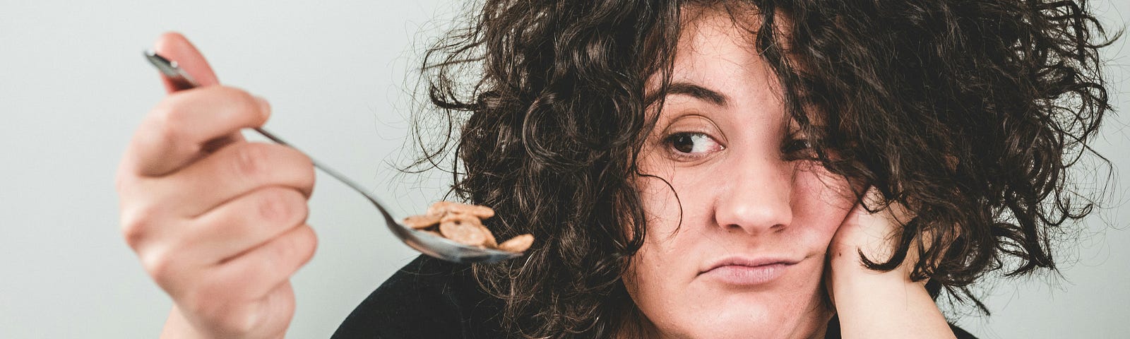 A woman with curly hair looks tired or bored as she holds a spoon over a bowl of cereal, resting her head on her hand.