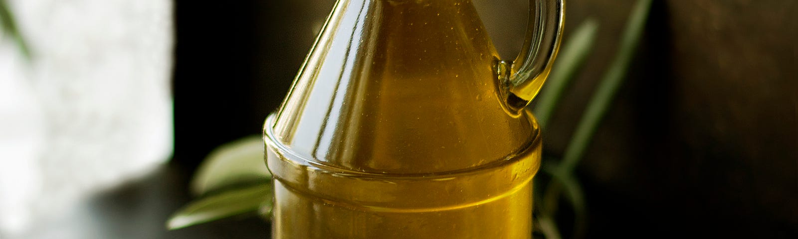 A glass bottle of olive oil sits on a bench surrounded by olives and herbs