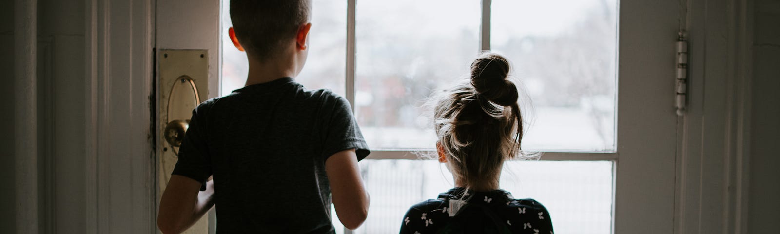 A young brother and still younger sister are standing side by side looking out of the window into a certain distance.