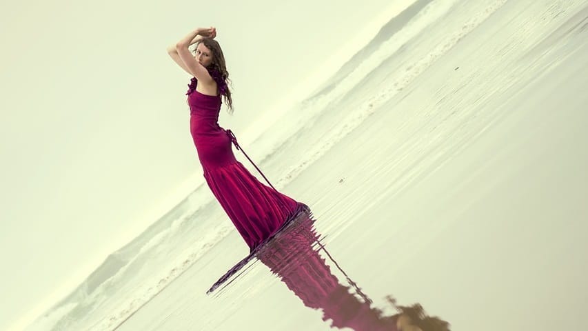 Woman in red dress standing on the beach, her hands over her head as if holding up the world.