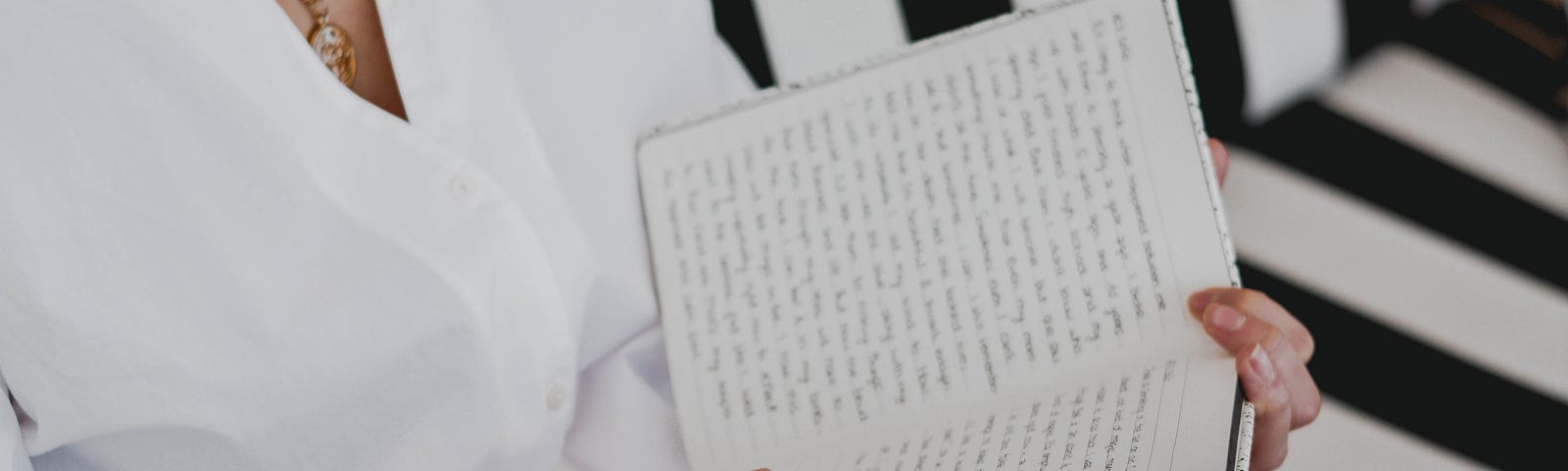 A woman in a white robe, sitting on a striped couch, writing in her notebook with a white pen.