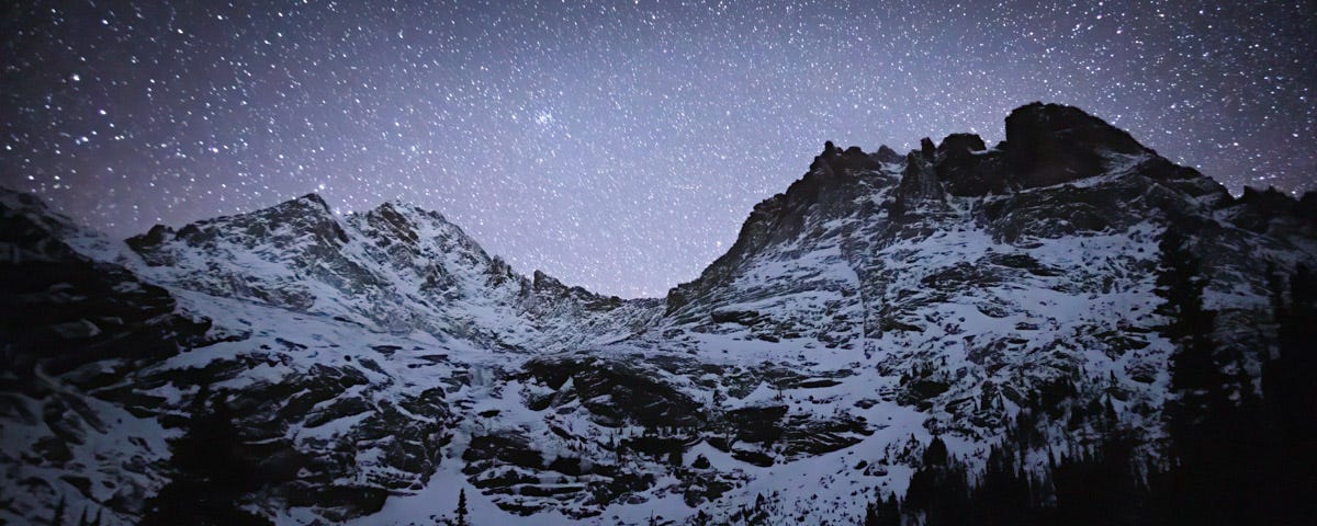 Black silhouetted trees form a base under rugged, snowy mountains, with an array of stars above.