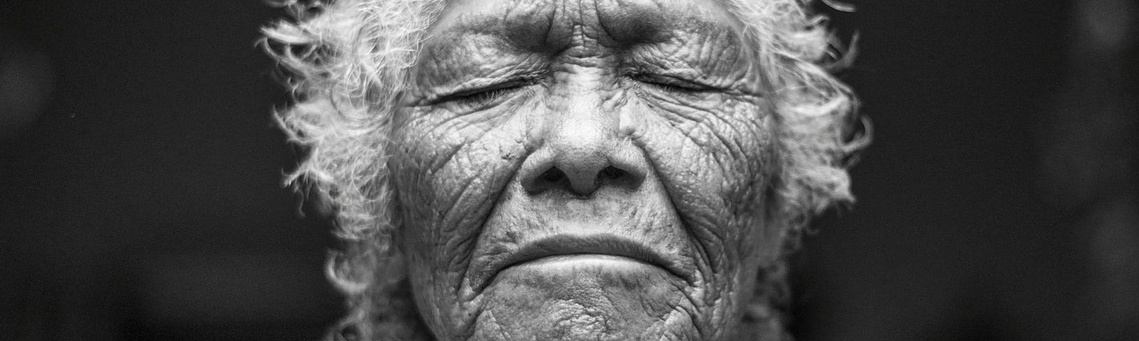 Photo of an old woman with white hair and brown skin. Countless lines decorate and accentuate her face, wrinkles of wisdom and time. Her eyes are closed, face tilted slightly up. Her lips are closed, and she appears to be listening to something deeper than sounds. She hears the wisdom and knowledge of the universe.