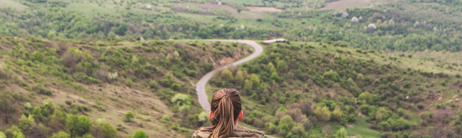 back of a female sitting on a rock overlooking a green, hilly landscape with a narrow road winding through it