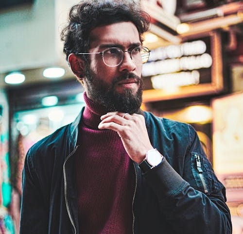 Stylish man in mauve turtleneck and black leather jacket in a well trimmed beard and glasses looking thoughtfully off to the side with a street scene in the background