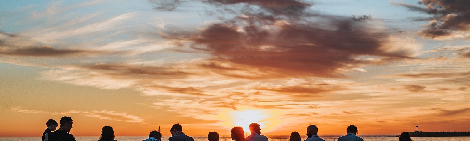 Image shows a large group of people, suggestive of a family, all holding hands or hugging, standing on the beach at sunset.