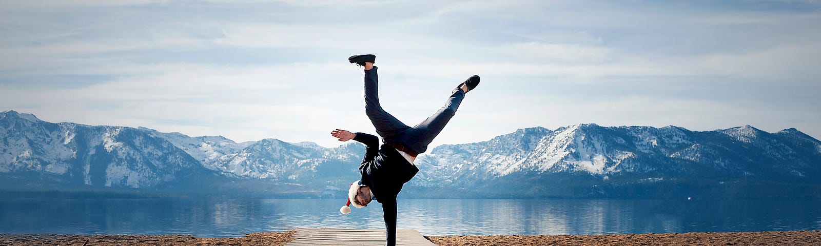 a man balancing on one hand on a long wooden walk way to a lake with mountains in the background